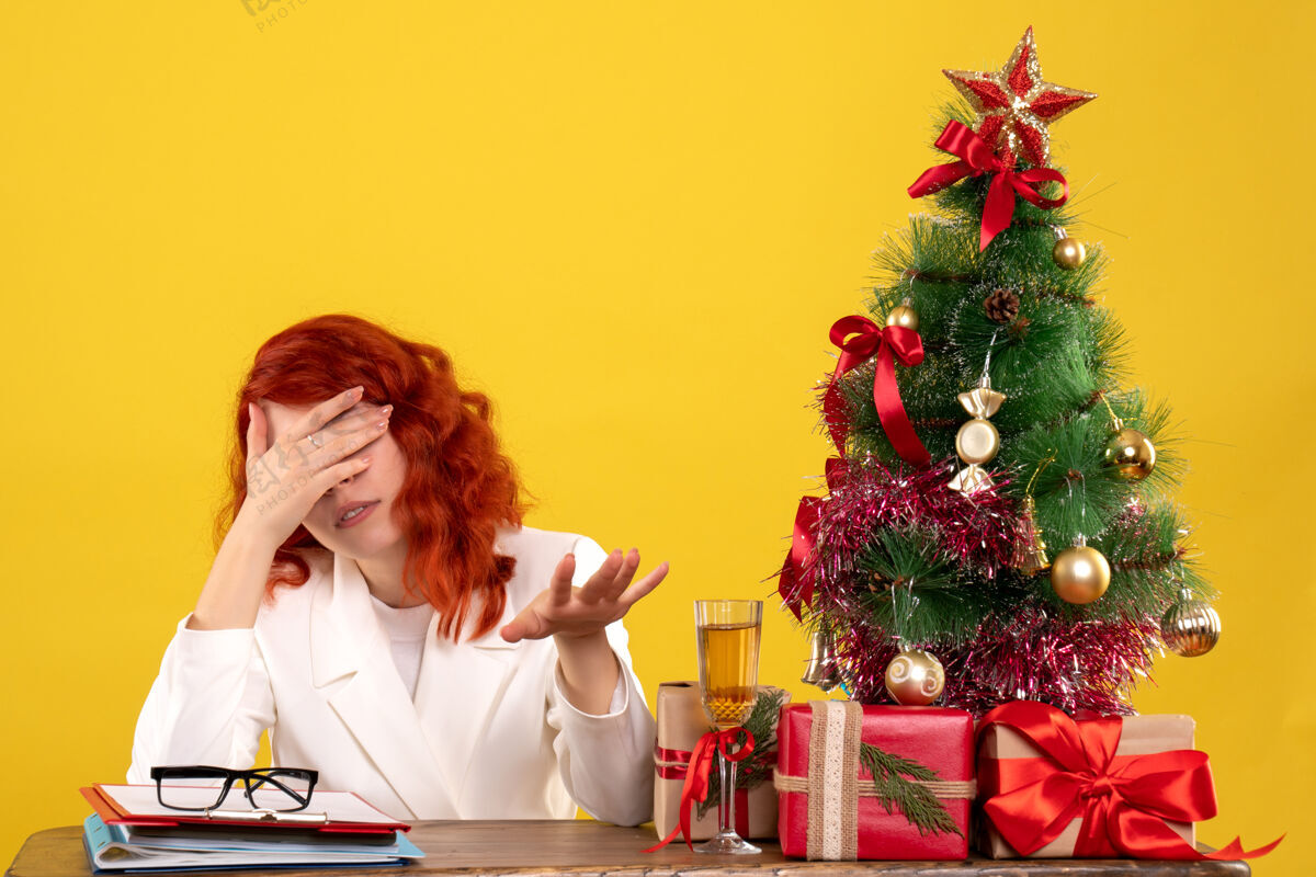 工人女工人坐在桌子后面 手里拿着圣诞礼物 圣诞树压在黄色上紧张桌子黄色