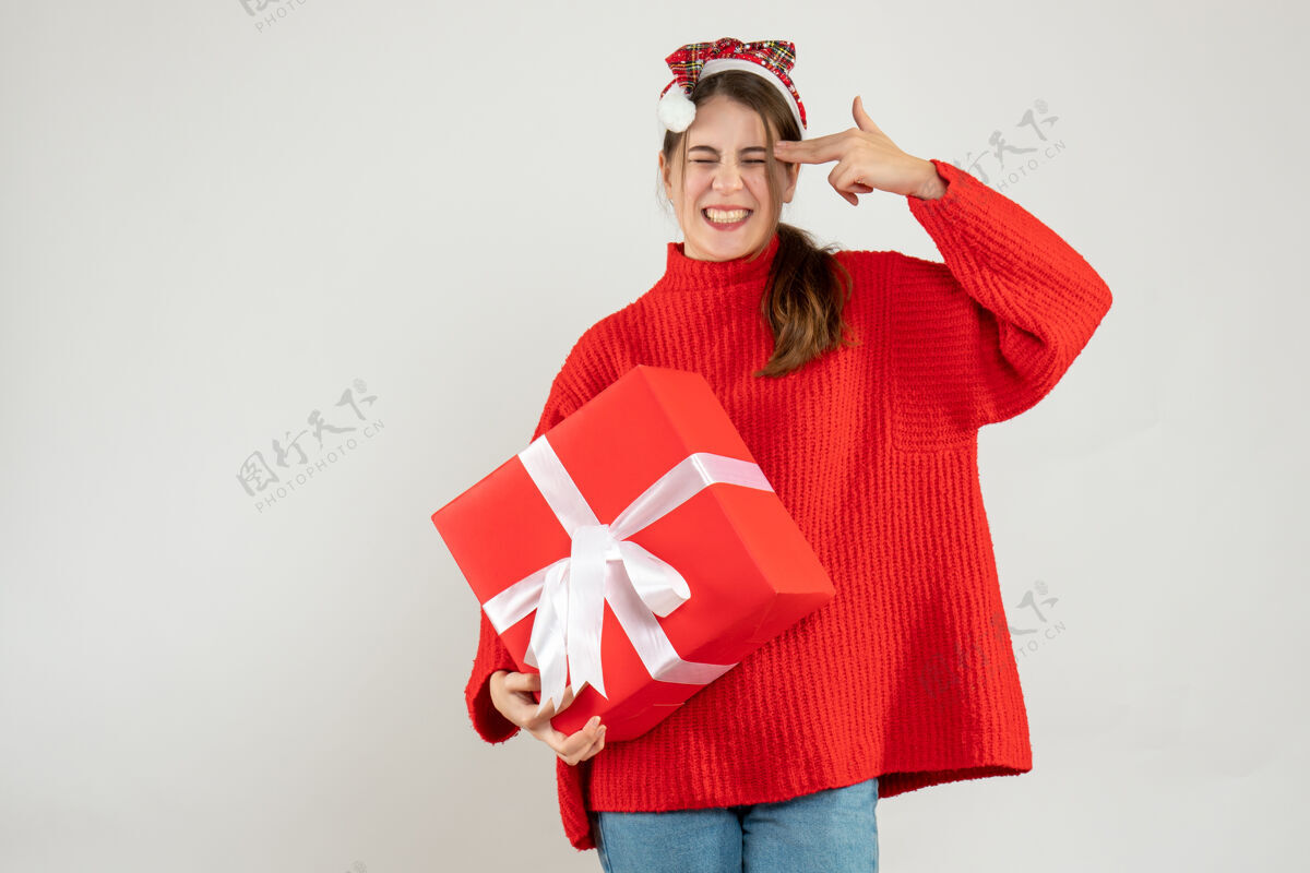 圣诞节戴着圣诞帽的快乐女孩拿着礼物把手指枪放在她的太阳穴上快乐举行女士