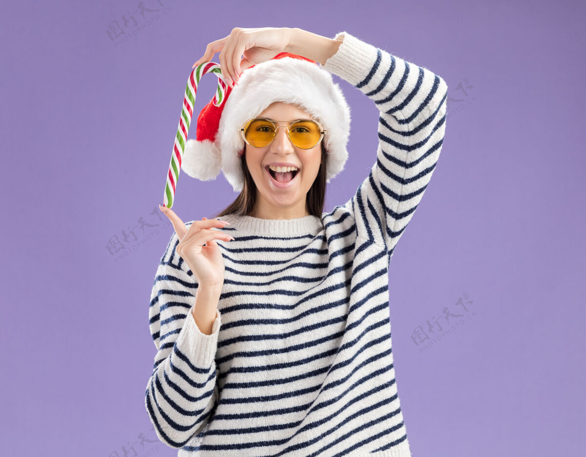 新快乐的年轻白人女孩戴着太阳眼镜 戴着圣诞帽 手里拿着一根糖果棒 在紫色的背景上与复制空间隔离开来快乐圣诞老人圣诞节