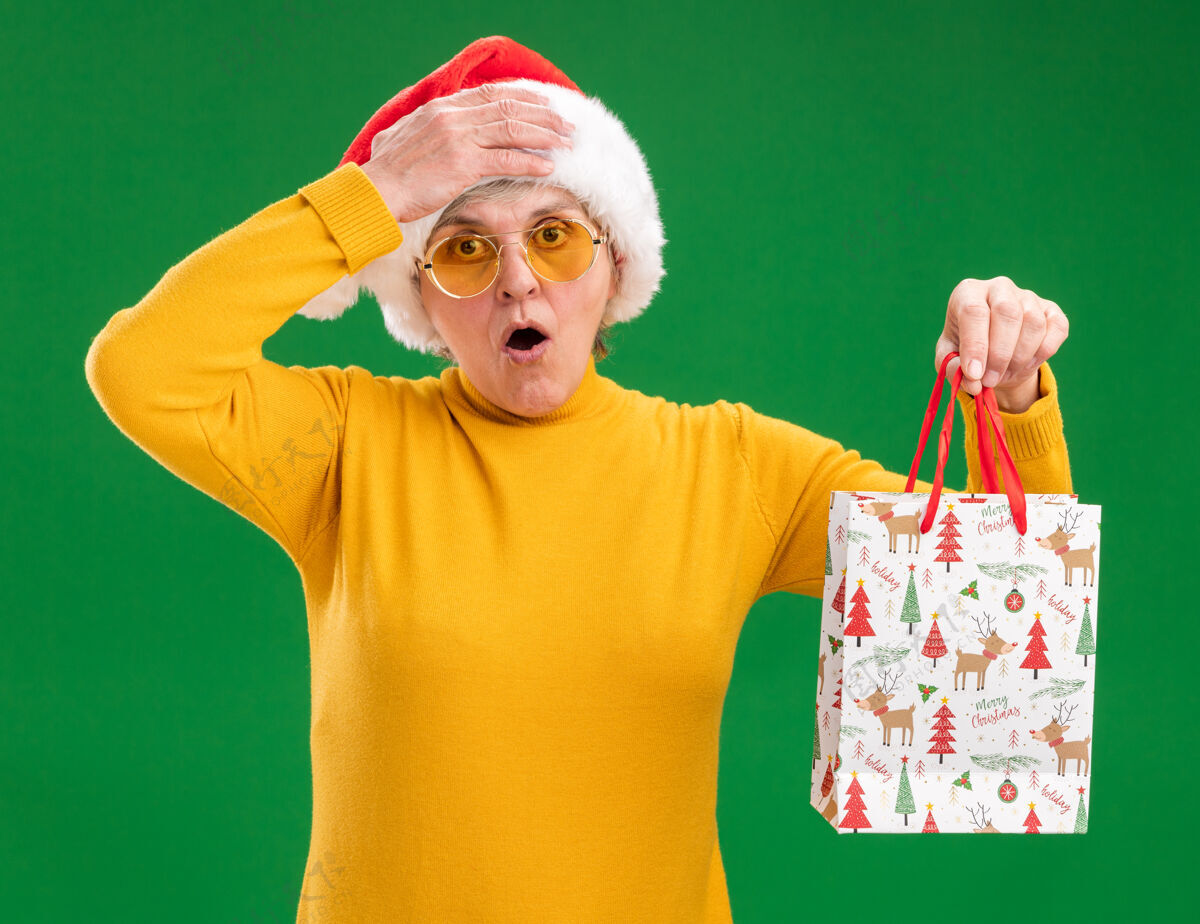 纸戴着太阳眼镜 戴着圣诞帽的老妇人吃惊地把手放在额头上 手里拿着纸礼包礼物空间帽子