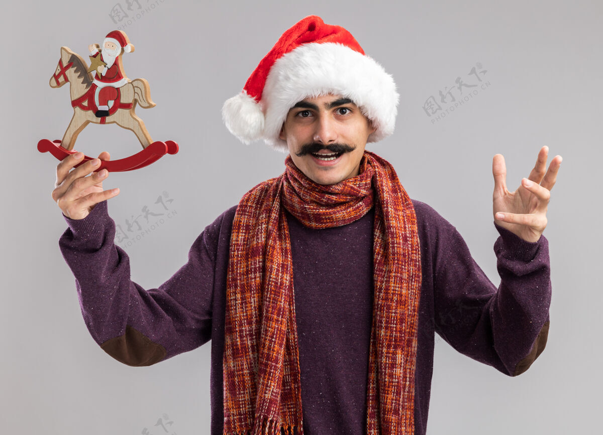 圣诞快乐年轻的胡子男人戴着圣诞老人帽 脖子上围着暖和的围巾 手里拿着圣诞玩具 兴高采烈地看着相机 举起胳膊站在白色的背景上周围胡子帽子