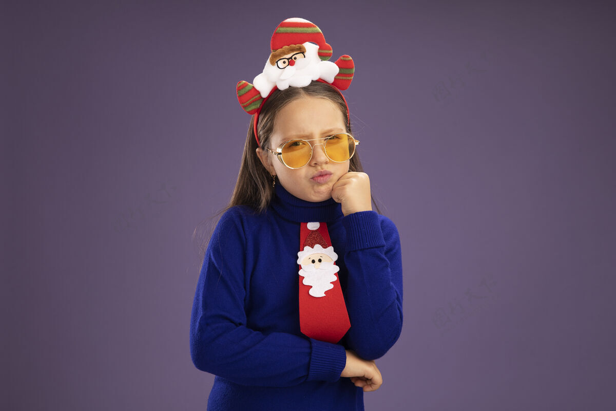 边缘小女孩穿着蓝色高领毛衣 打着红色领带 头上戴着滑稽的圣诞戒指 站在紫色的背景下 带着怀疑的表情看着相机圣诞节搞笑领带