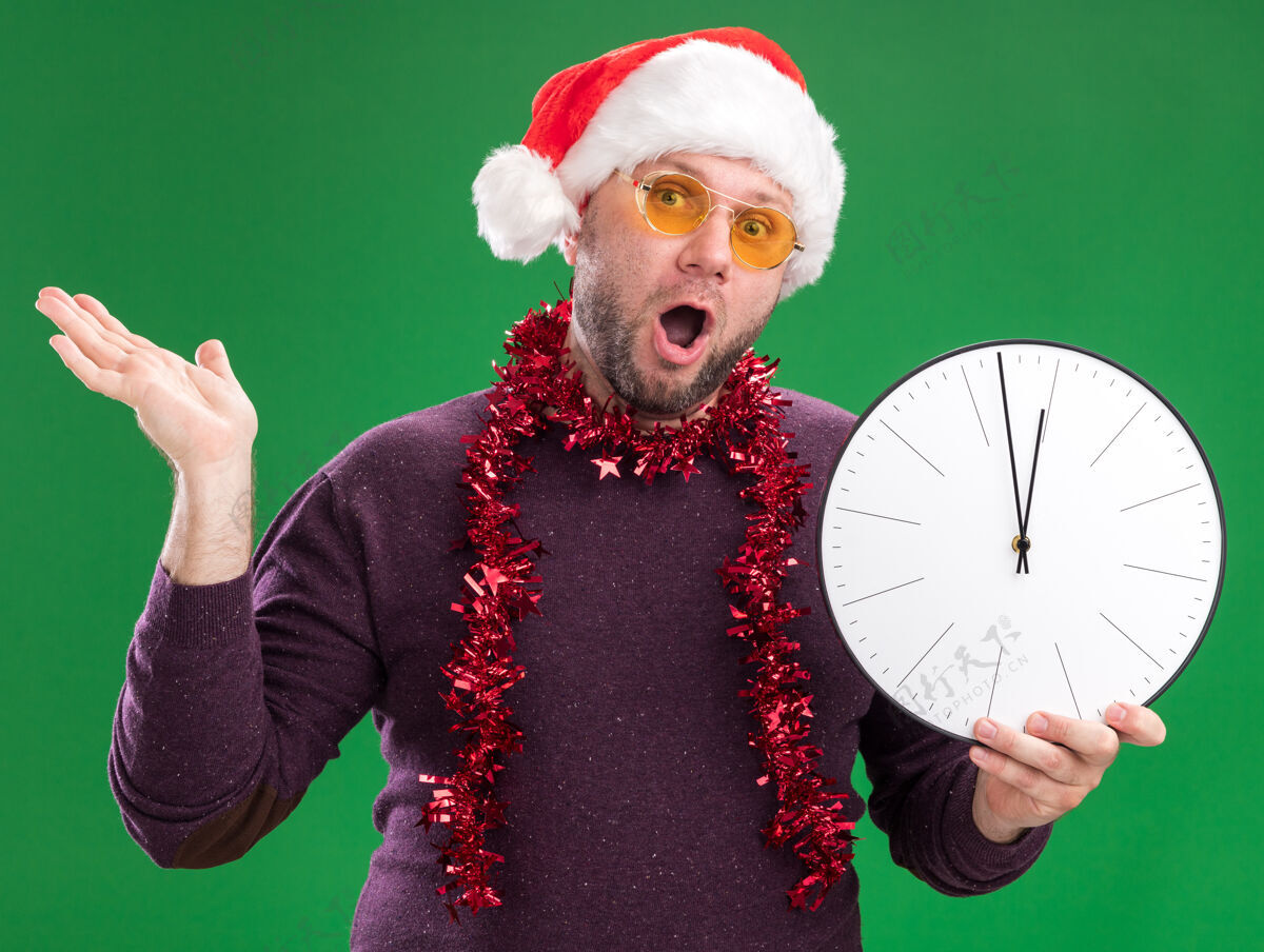 男人令人印象深刻的中年男子戴着圣诞老人帽 脖子上戴着金属丝花环 戴着眼镜 拿着一只被隔离在绿色墙上的空手的钟时钟金属片印象深刻