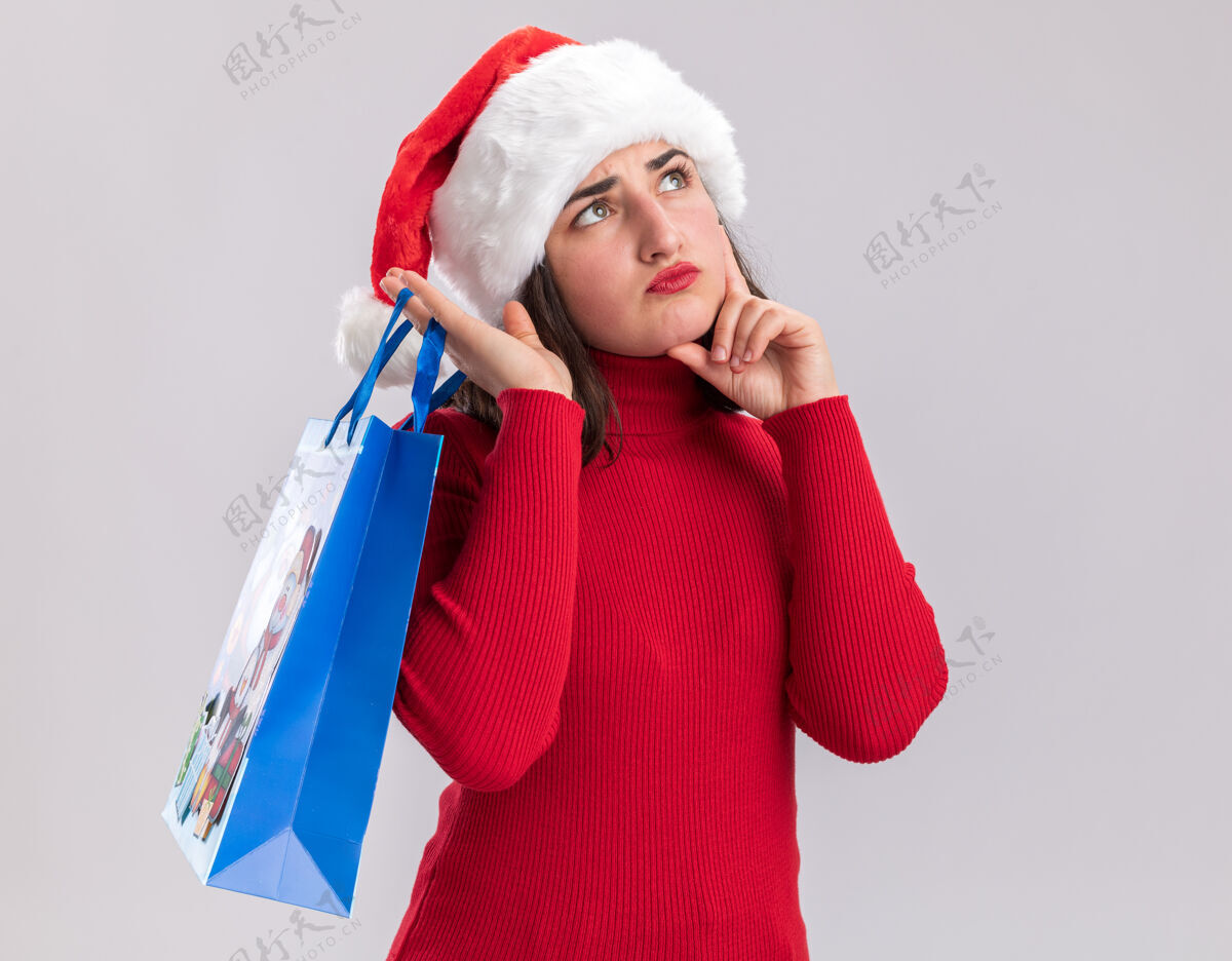 包穿着红色毛衣 戴着圣诞帽的小女孩 手里拿着五颜六色的圣诞礼物纸袋 带着沉思的表情抬起头 站在白色的背景上思考表情礼物抱着
