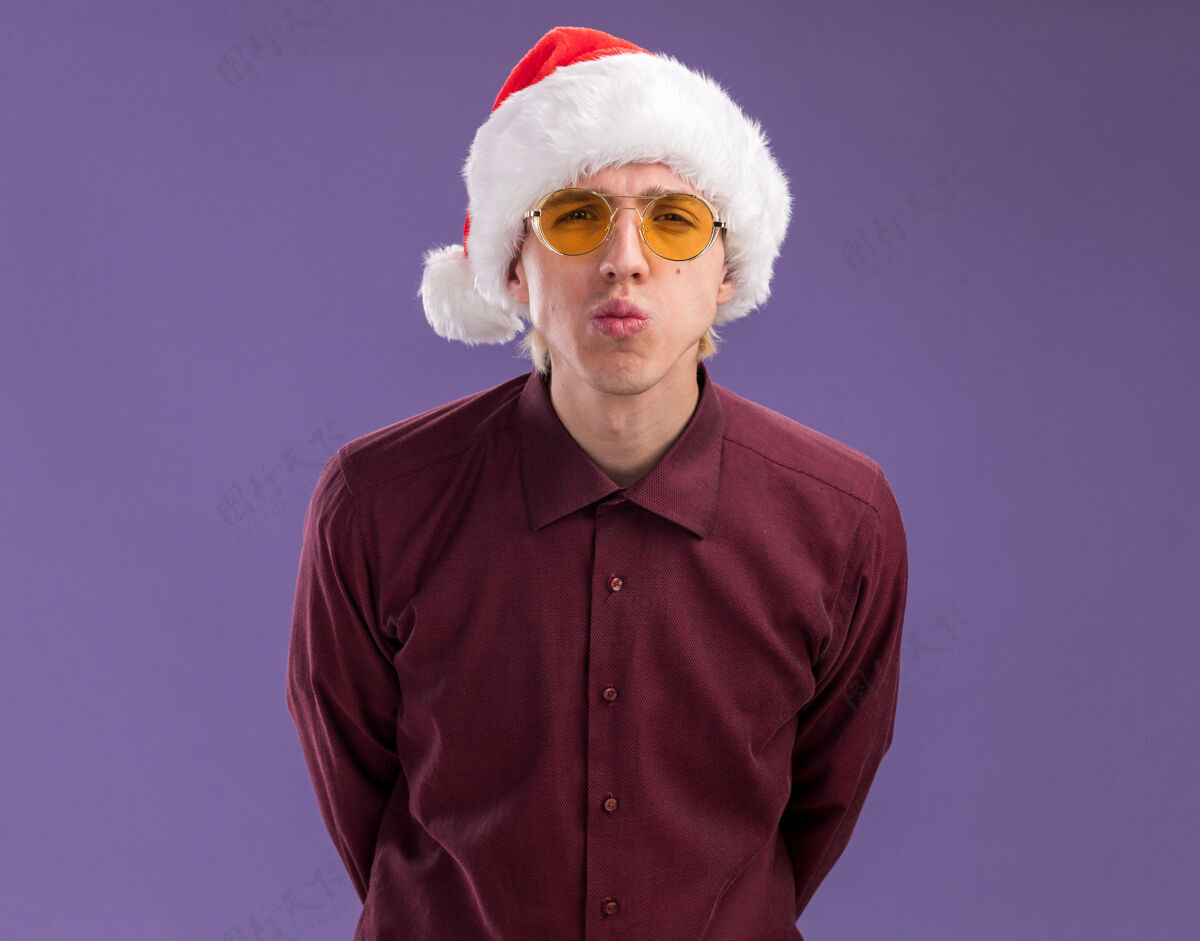 自信自信的年轻金发男子戴着圣诞老人的帽子和眼镜看着摄像机保持双手在背后做亲吻手势隔离紫色背景帽子保持金发