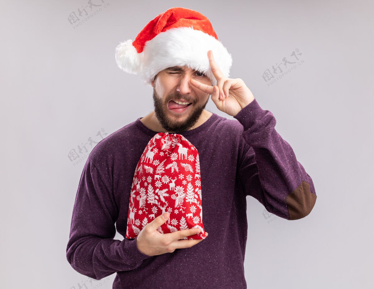 舌头一个穿着紫色毛衣 戴着圣诞老人帽的年轻人 手里拿着一个红色的礼物包 上面有一个v字标志 眼睛附近有一个突出的舌头 站在白色的背景上毛衣站着年轻人