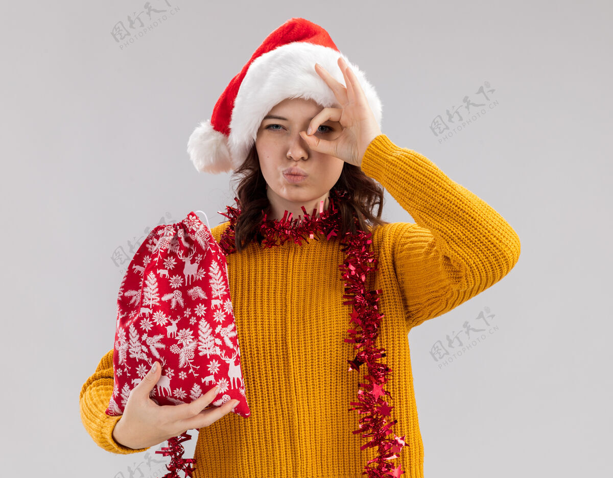 包自信的年轻斯拉夫女孩 戴着圣诞帽 脖子上戴着花环 手里拿着圣诞礼品袋 手指隔着白色背景和复印空间看着相机举行花环礼物