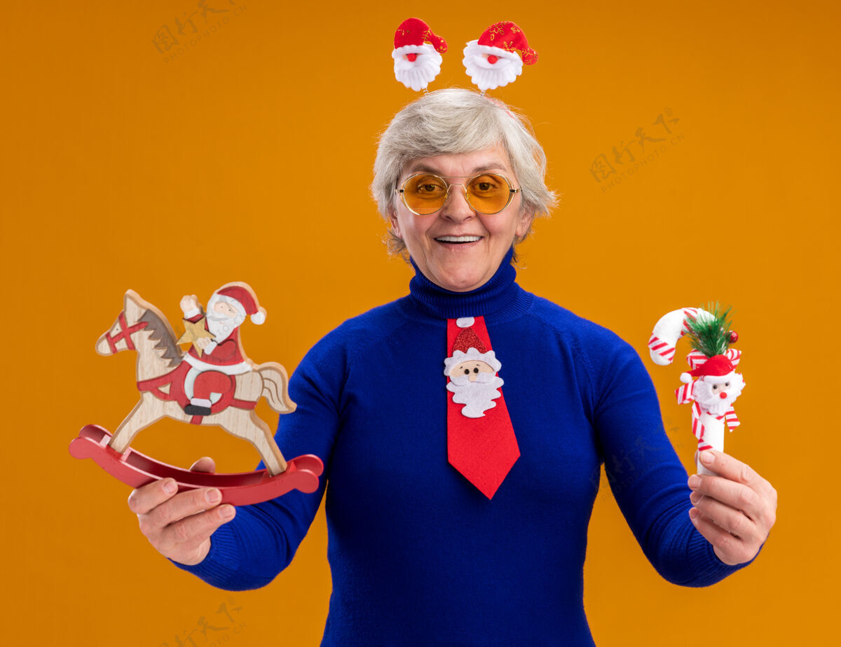 圣诞老人戴着太阳眼镜微笑的老妇人 戴着圣诞老人头带 打着圣诞老人领带 把圣诞老人抱在摇摇木马上 把糖果手杖隔离在橙色背景上 留有复制空间头带微笑眼镜