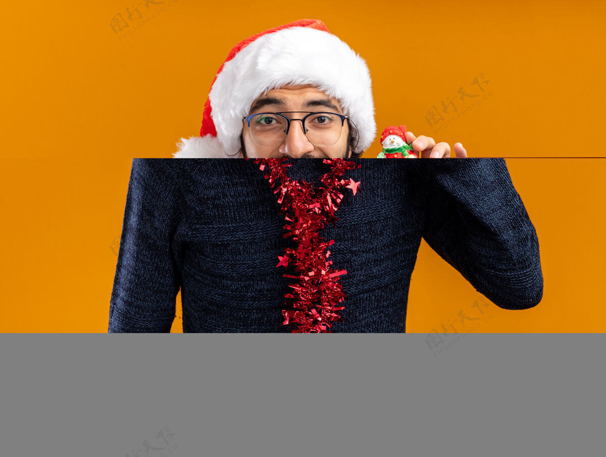 花环兴奋的年轻帅哥戴着圣诞帽 脖子上戴着花环 手里拿着橙色墙上孤立的玩具脖子帽子年轻