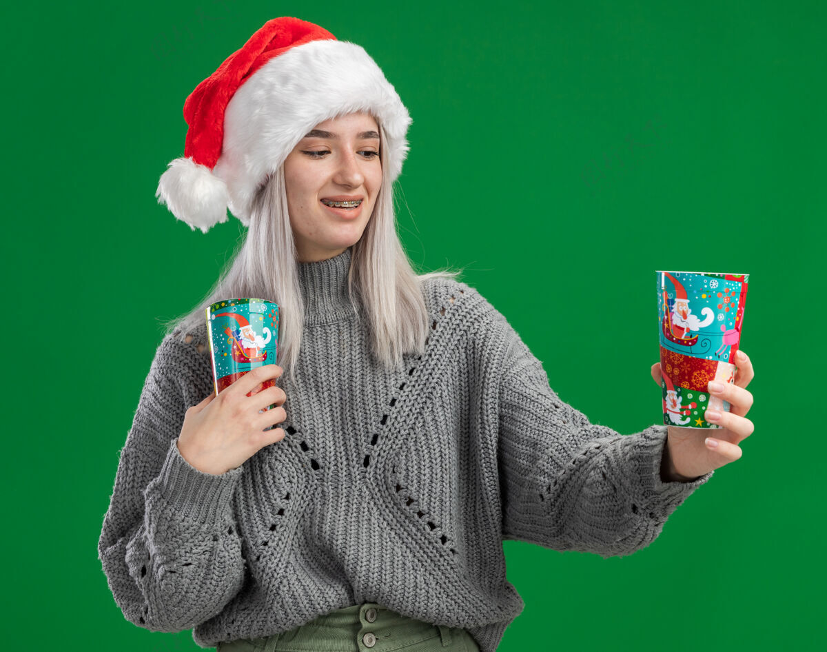 杯子年轻的金发女郎穿着冬衣 戴着圣诞帽 手里拿着五颜六色的纸杯 站在绿色的背景下 面带微笑地看着杯子杯子站着毛衣