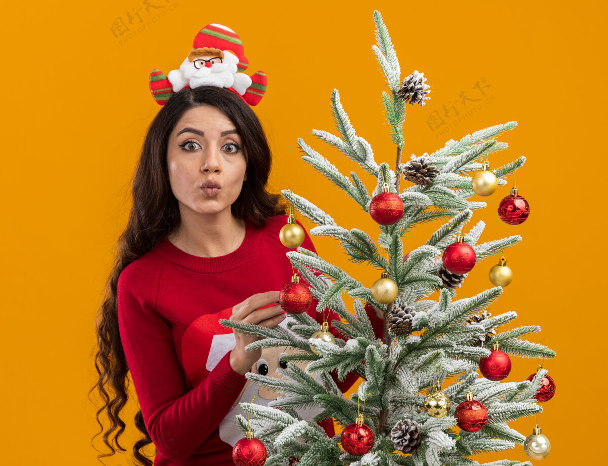 圣诞树给人印象深刻的年轻漂亮女孩戴着圣诞老人的头带和毛衣站在装饰好的圣诞树后面看着相机噘着嘴唇孤立在橙色背景上圣诞年轻印象深刻