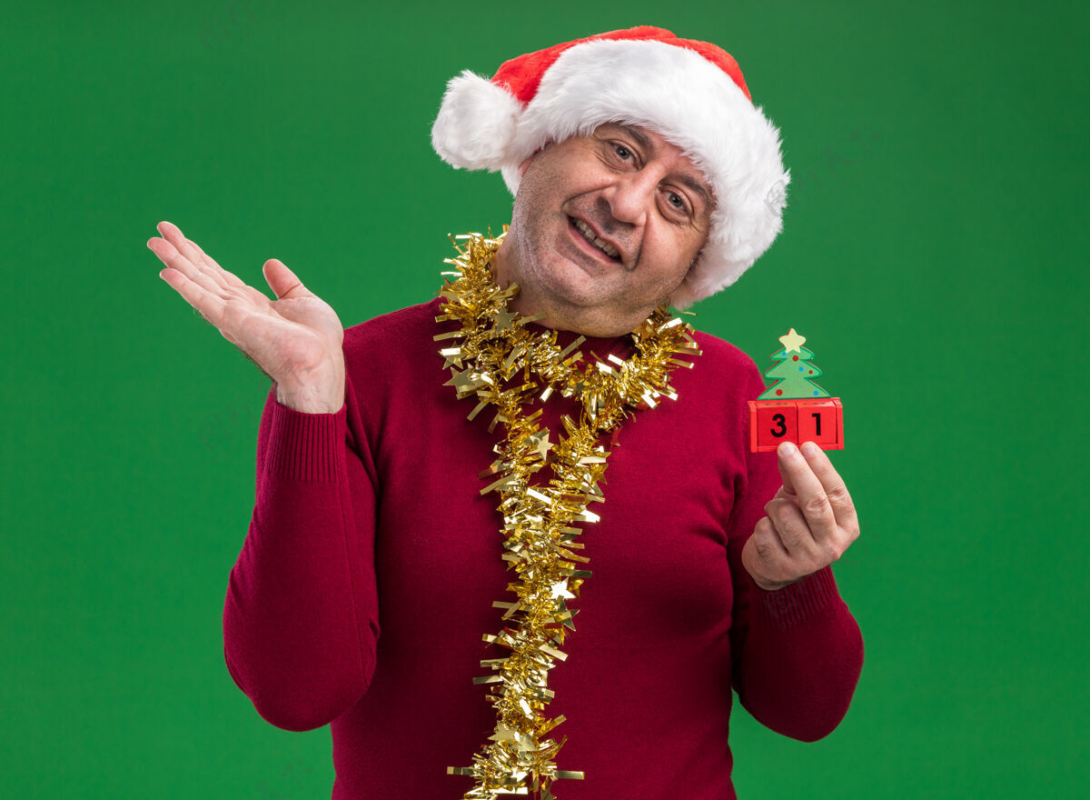 站着中年男子戴着圣诞老人帽子 脖子上戴着金属丝 手里拿着玩具方块 手里拿着新年的日期 看着相机微笑着 开心地抬起手臂站在绿色的背景上新的抱着手臂