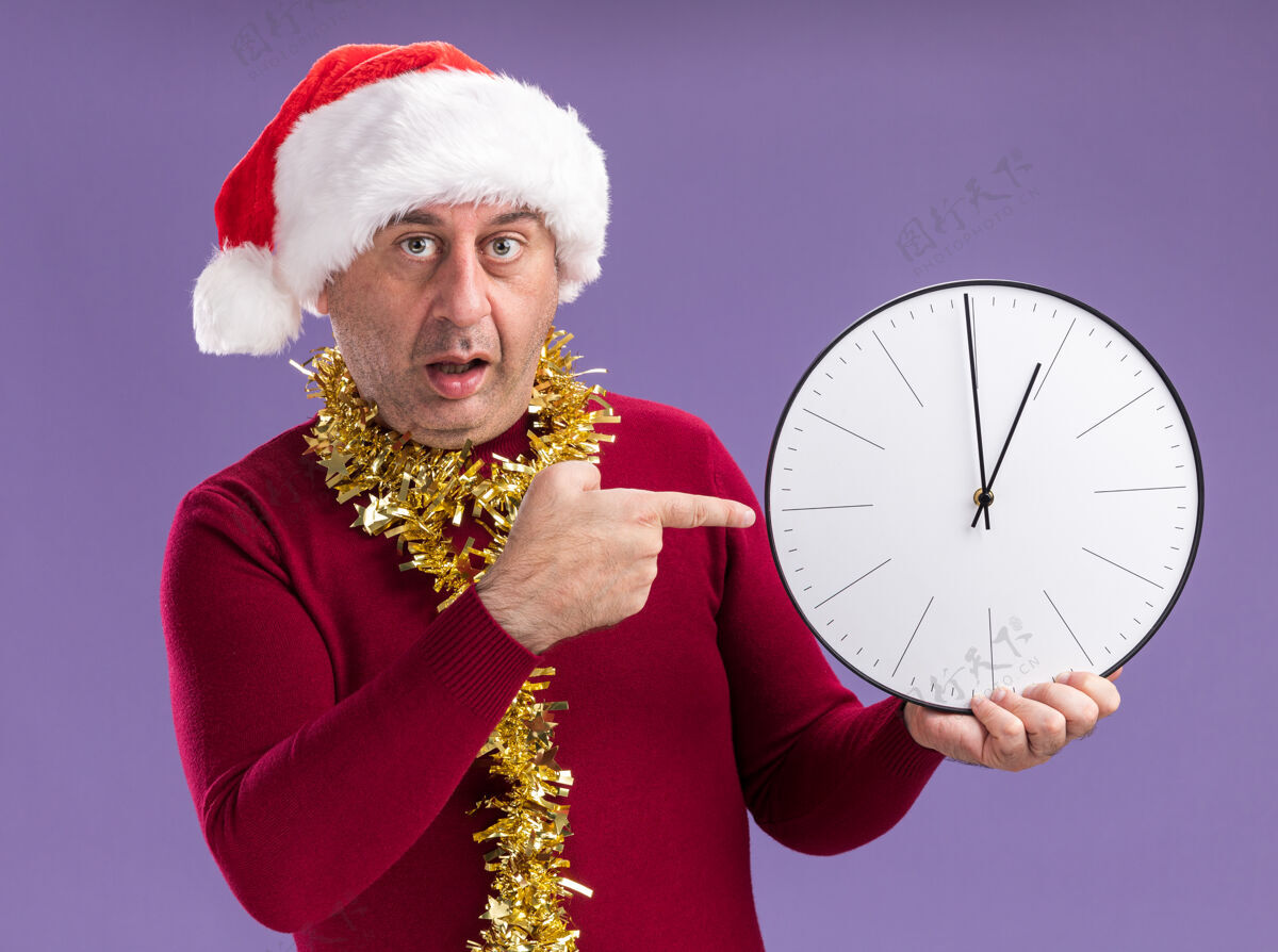 拿着中年男子戴着圣诞老人的帽子 脖子上戴着金属丝 拿着钟 用食指指着它 站在紫色的背景下 神情忧虑脖子担心中间