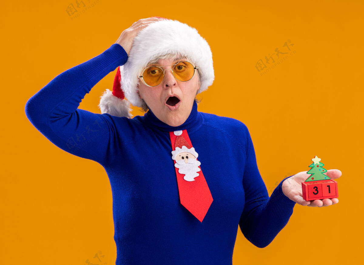圣诞戴着太阳眼镜 戴着圣诞帽 打着圣诞领带的惊讶的老妇人拿着圣诞树饰物 把手放在孤立的橙色背景上 留有复制空间橙色头惊喜