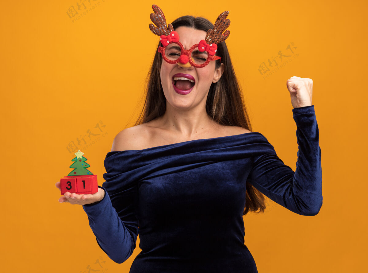 手势兴奋地闭上眼睛年轻漂亮的女孩穿着蓝色的裙子和圣诞眼镜举行玩具显示是的姿态孤立在橙色的墙上展示服装穿着
