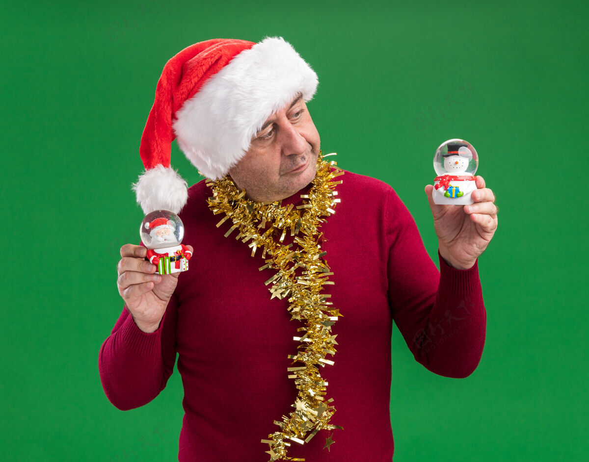 地球仪中年男子戴着圣诞老人帽 脖子上戴着金属丝 手里拿着圣诞雪球 站在绿色的背景上 看上去很困惑 有疑虑举行脖子帽子