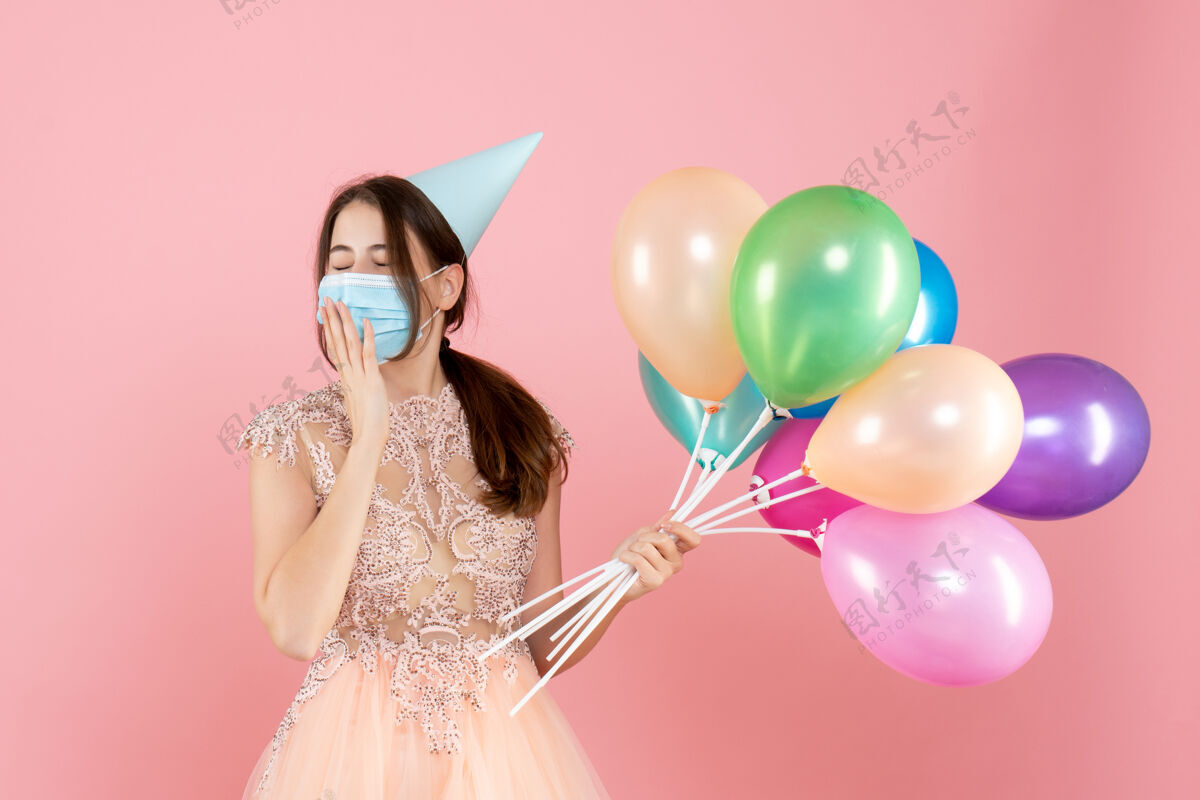举行睡意朦胧的女孩 戴着派对帽和医用面具 手里拿着粉色的彩色气球粉红色气球生日