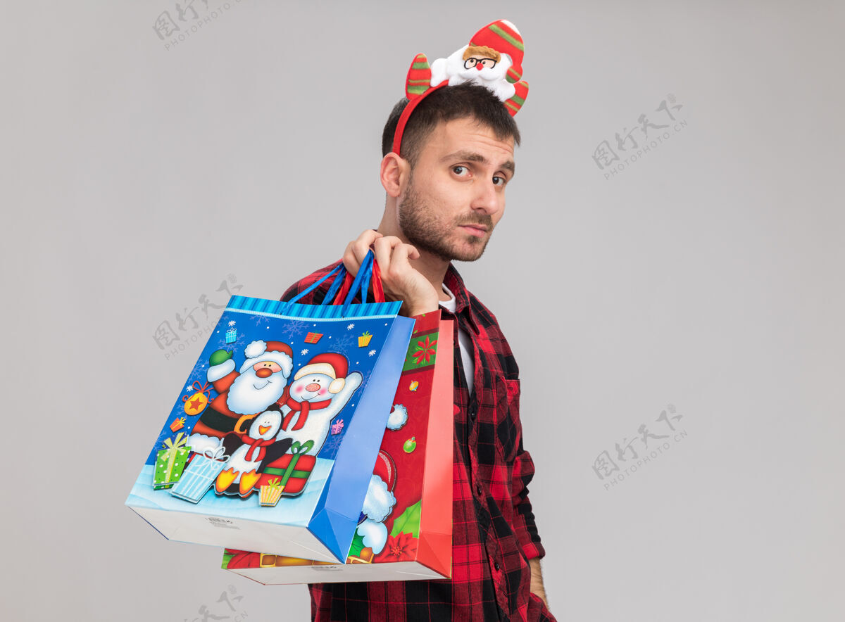 头带自信的白人年轻人戴着圣诞头带站在侧视图中 肩上扛着圣诞礼品袋 看着隔离在白色背景上的相机 还有复印空间个人资料圣诞快乐礼物