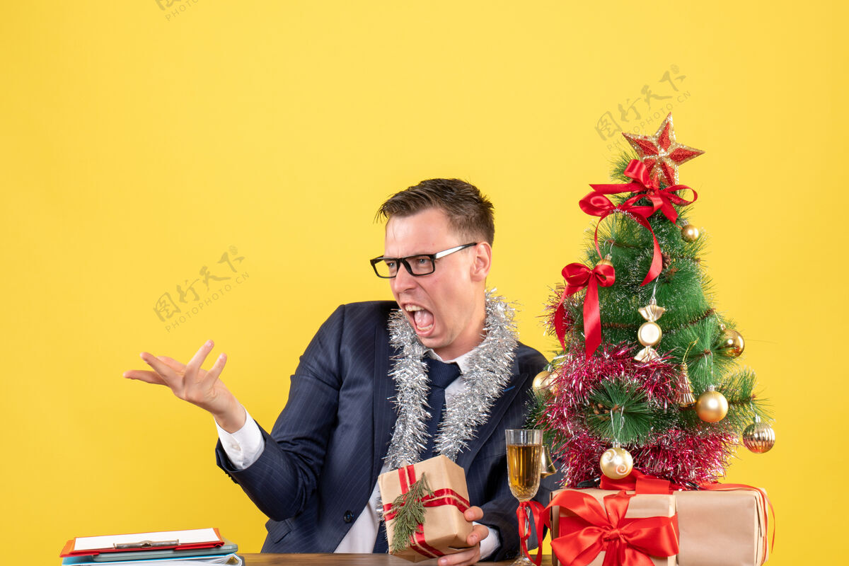 微笑愤怒的男人戴着眼镜坐在圣诞树旁的桌子前 黄色的墙上摆着礼物成人人人