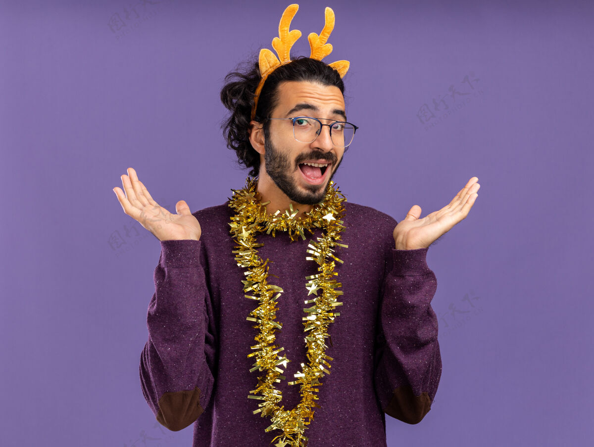 圣诞节快乐的年轻帅哥戴着圣诞发箍 脖子上戴着花环 双手分开放在蓝色的墙上年轻快乐箍