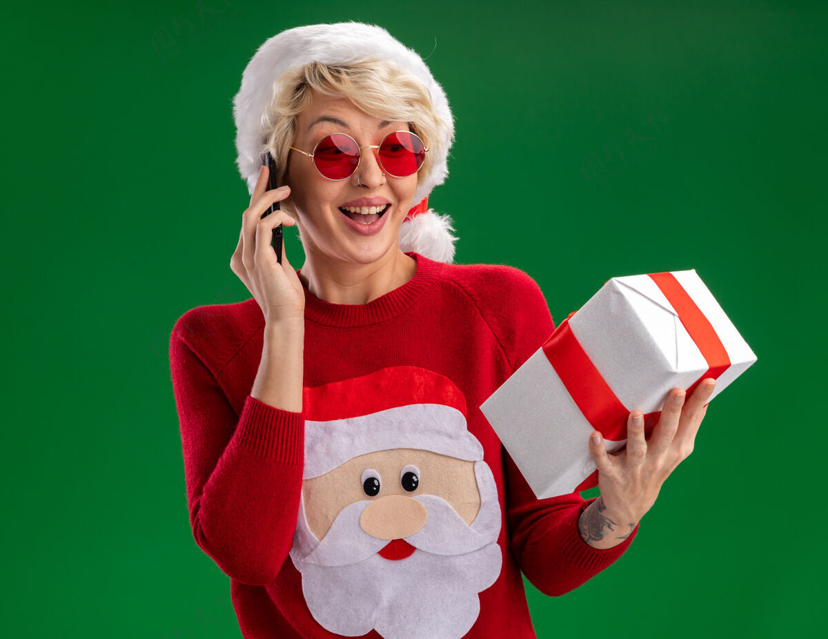 圣诞节快乐的金发女郎戴着圣诞帽 穿着圣诞老人的圣诞毛衣 戴着眼镜 拿着礼物包 隔着绿色的墙壁在电话里聊天包装快乐女人
