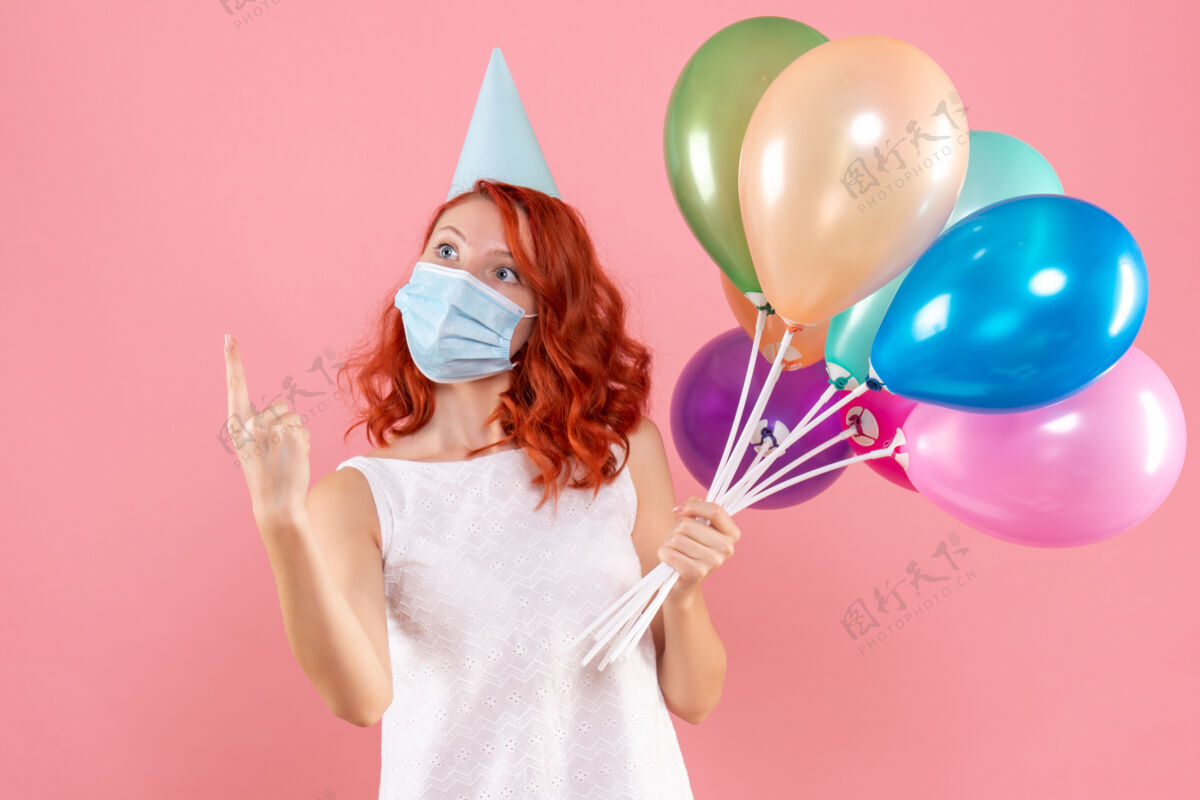 气球前视图中的年轻女子拿着五颜六色的气球在无菌面具上粉红色的墙壁无菌聚会快乐