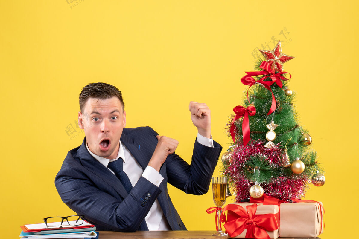 桌子前视图惊讶的男子手指指着后面坐在桌旁圣诞树和礼物上的黄色墙壁团队手指礼物