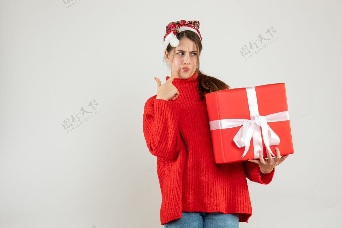 放体贴的女孩戴着圣诞帽把手指放在脸颊上拿着白色的礼物漂亮抱脸颊