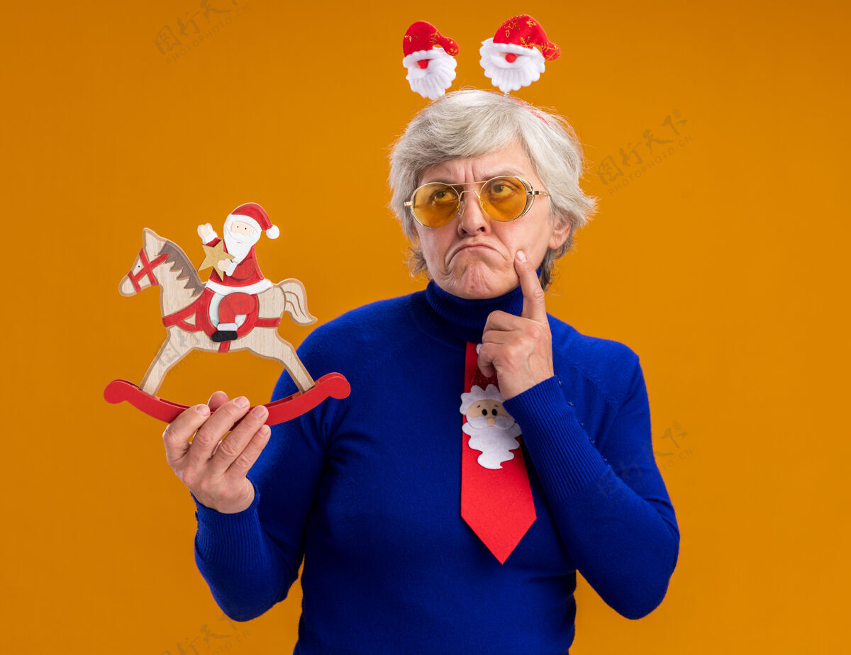 头带戴着太阳眼镜 戴着圣诞老人头带和圣诞老人领带的体贴的老妇人把手指放在下巴上 把圣诞老人抱在摇马装饰上 在橙色的背景上孤立地向上看 还有复制空间下巴圣诞老人体贴