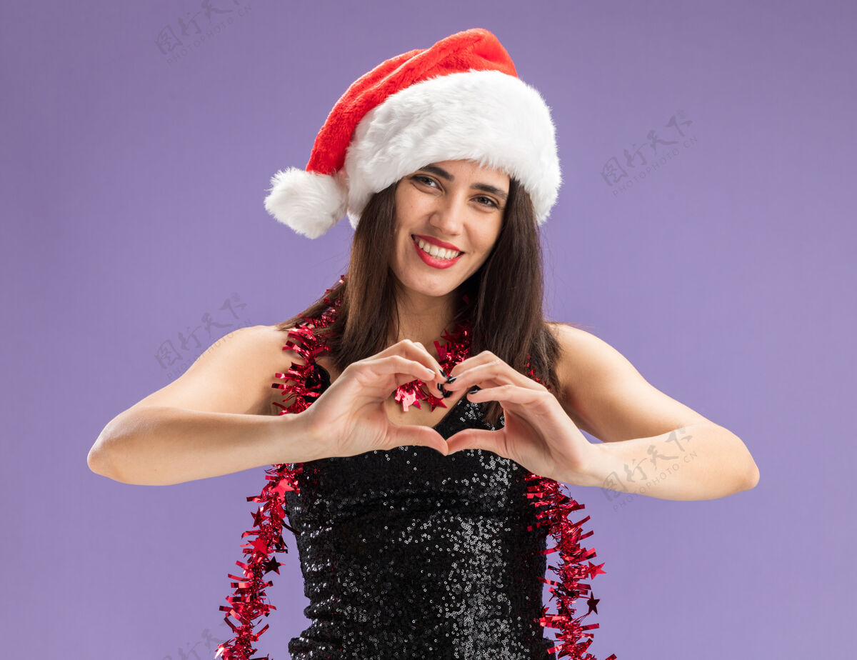 女孩微笑的年轻漂亮女孩戴着圣诞帽 脖子上戴着花环 在紫色的背景上显示出孤立的心形花环圣诞节帽子