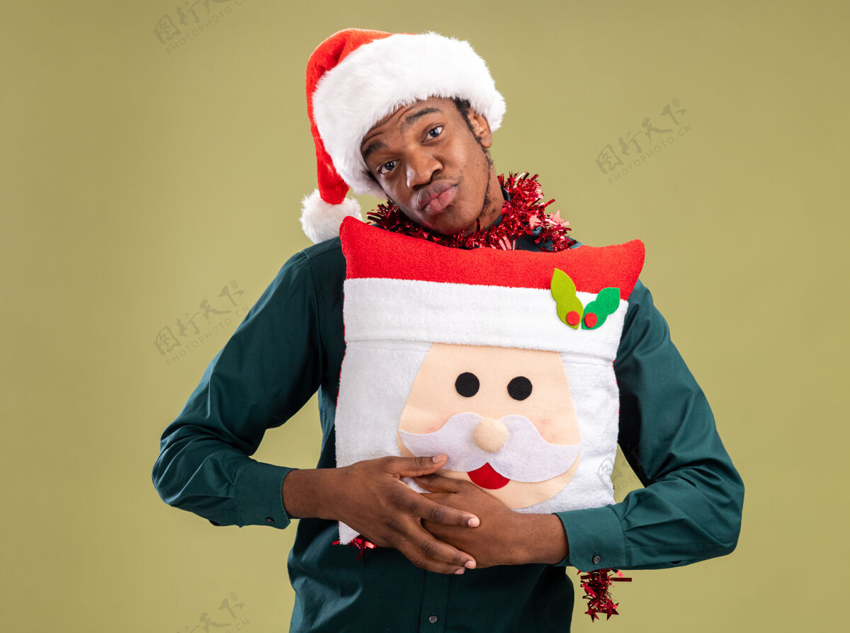 花环非洲裔美国人戴着圣诞帽 戴着花环 抱着圣诞枕头 站在绿色的背景下 快乐而积极地看着镜头圣诞节枕头举行