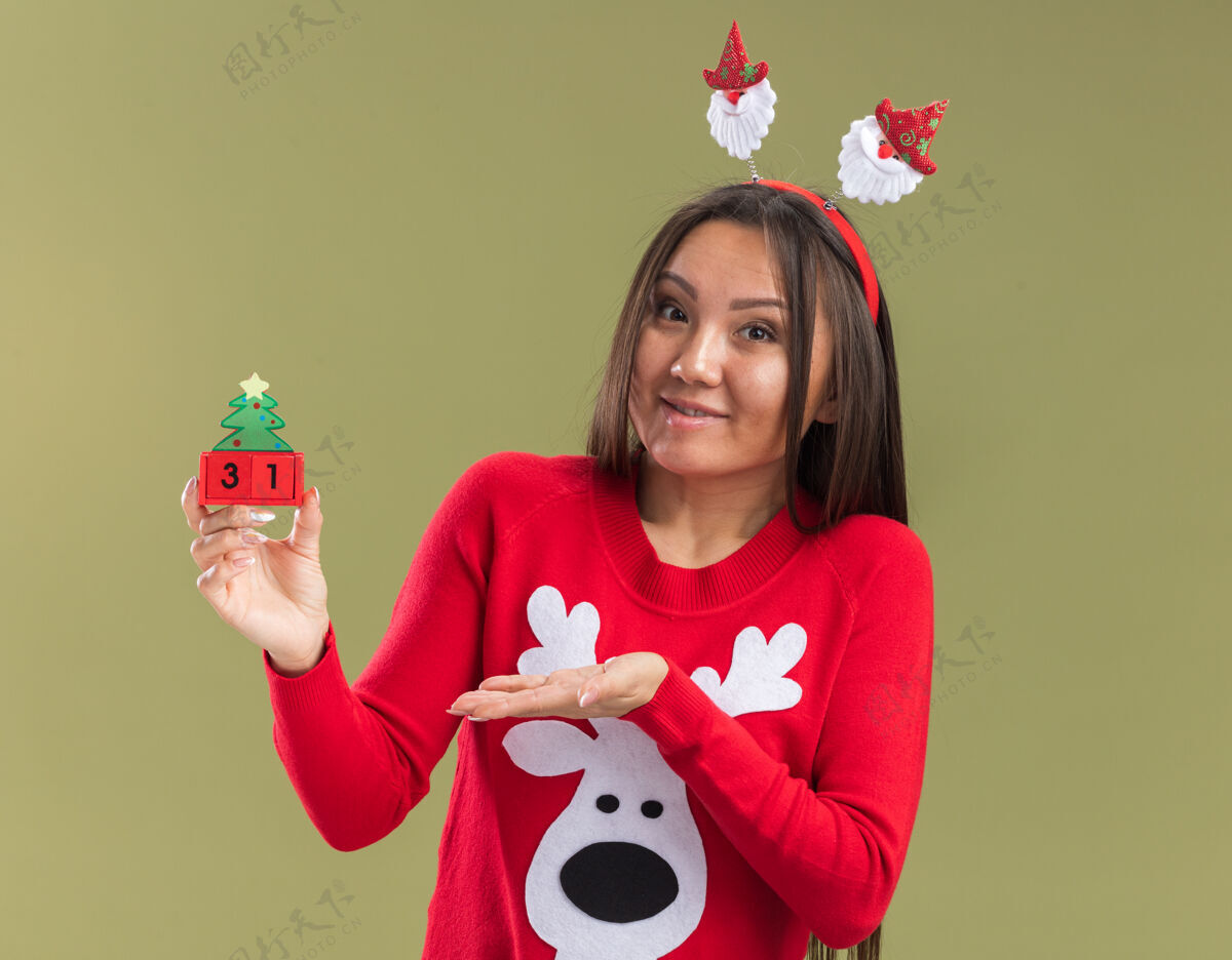 高兴高兴的年轻亚洲女孩戴着圣诞发箍抱着和手指在圣诞玩具孤立的橄榄绿背景头发抱女孩