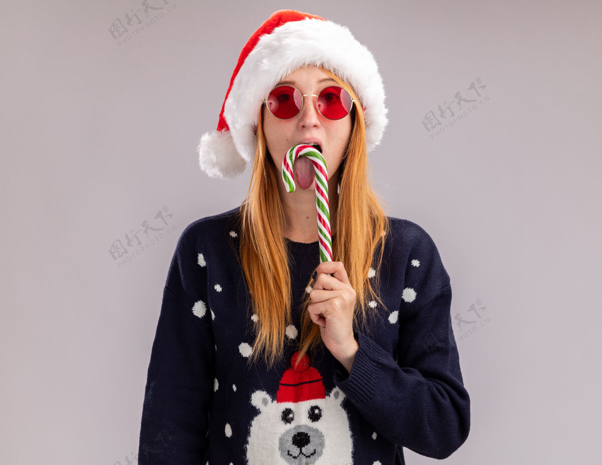 穿年轻漂亮的女孩戴着圣诞帽 戴着眼镜 抱着和舔着隔离在白墙上的圣诞糖果糖果舔圣诞节