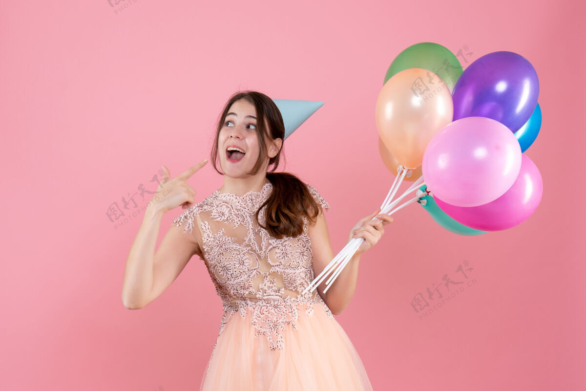 帽子快乐的派对女孩 戴着派对帽 拿着气球 手指指着粉色的脸笑快乐的聚会女孩气球快乐