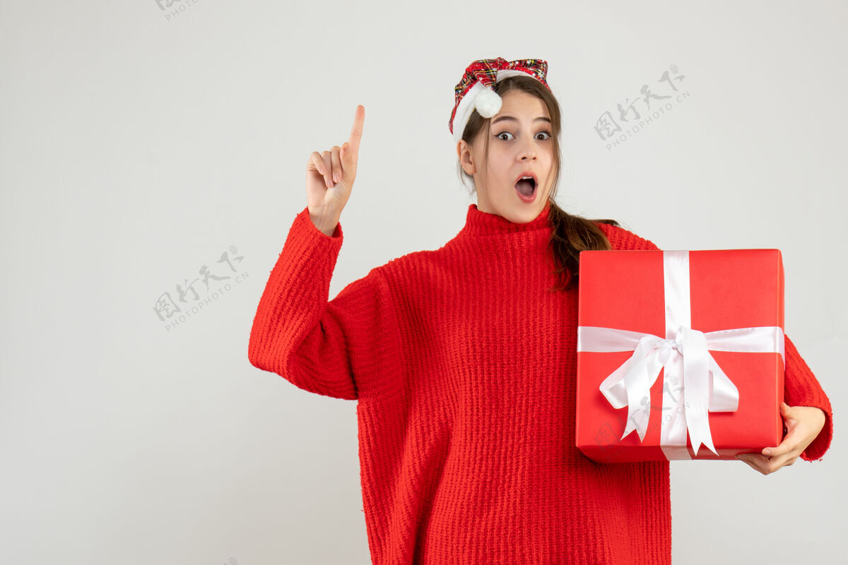 圣诞节戴着圣诞帽的好奇女孩拿着礼物手指着站在白色地板上拿着女孩好奇