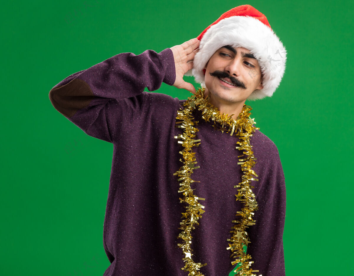 金属片年轻的胡子男人戴着圣诞老人帽子 脖子上挂着金属丝 站在绿色的背景下 面带微笑地看着一边圣诞节脖子脸