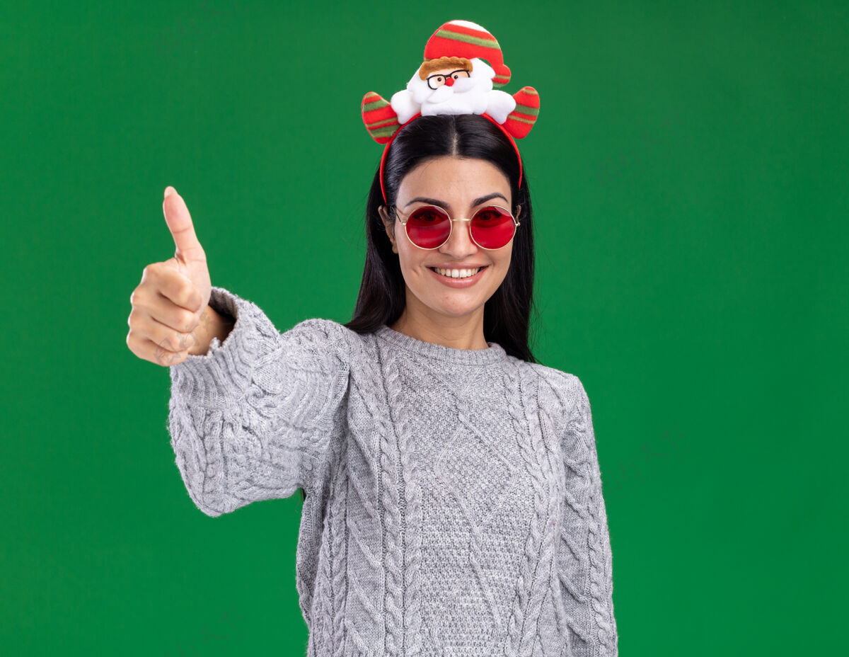 圣诞老人戴着圣诞老人头带 戴着眼镜 微笑着的年轻白人女孩看着相机 在绿色背景上孤立地竖起大拇指显示拇指微笑