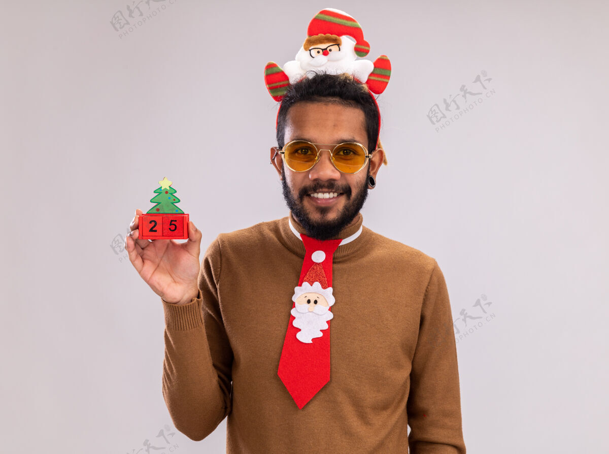 非洲快乐的非洲裔美国人 穿着棕色毛衣 头戴圣诞老人圈 打着有趣的红色领带 手里拿着玩具方块和25岁的约会对象 站在白色背景下微笑着看着相机搞笑立方体举行