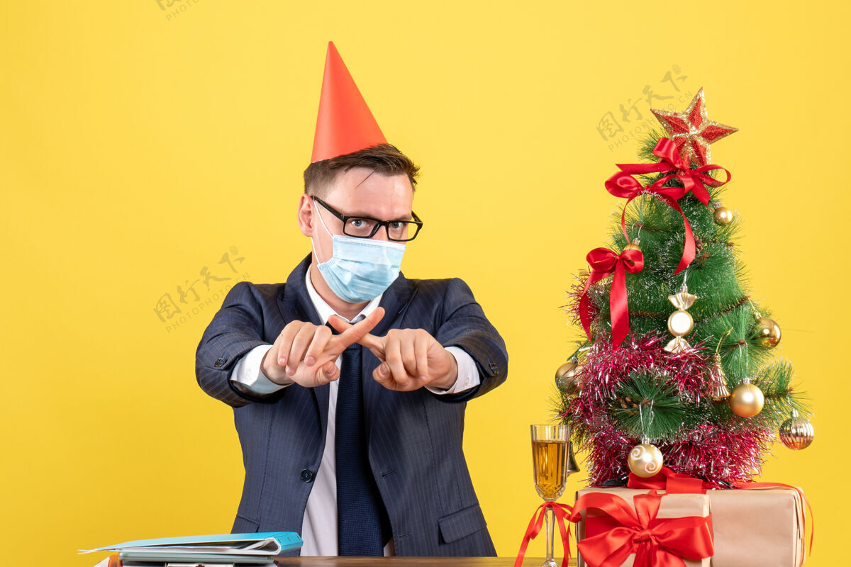坐商务人士交叉手指坐在圣诞树旁的桌子前 黄色墙壁上的礼物新正常人冠状病毒