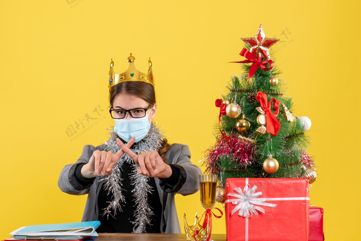 十字架前视图：戴着医用面罩的年轻女孩交叉手指 圣诞树和鸡尾酒礼物面具桌子医学