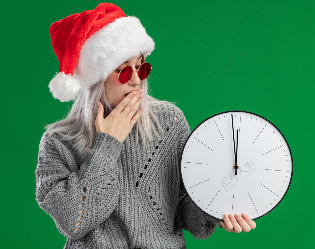 封面年轻的金发女郎穿着冬季毛衣 戴着圣诞老人帽 戴着红眼镜 拿着挂钟看着它 惊讶地捂着嘴 手站在绿色的背景上圣诞老人毛衣眼镜