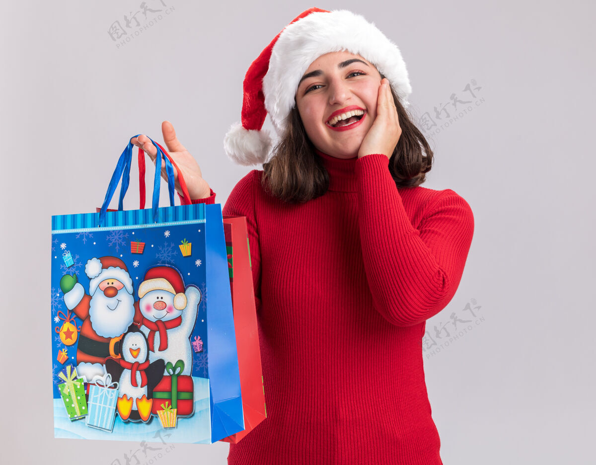 女孩穿着红色毛衣 戴着圣诞帽的小女孩手里拿着五颜六色的纸袋 手里拿着圣诞礼物 站在白色的背景上 满脸幸福地看着相机礼物年轻举行