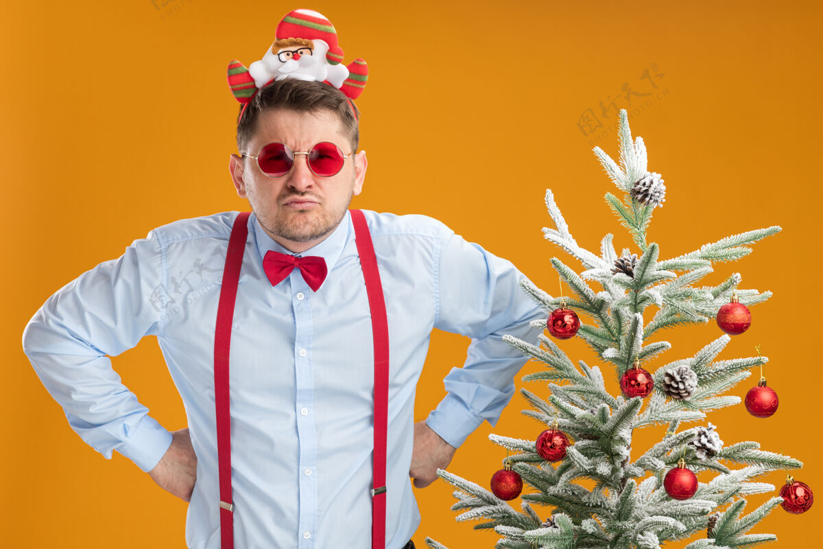 领带不高兴的年轻人穿着吊带领结与圣诞老人和红色眼镜站在旁边的圣诞树在橙色背景下愤怒的脸看着相机站着边缘年轻