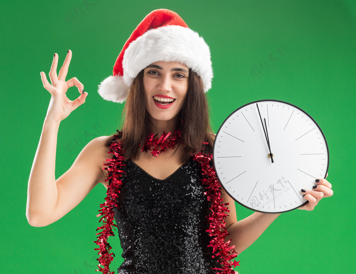 时钟微笑的年轻漂亮女孩戴着圣诞帽 脖子上戴着花环 手持挂钟 在绿色背景上显示出良好的姿态花环墙壁帽子