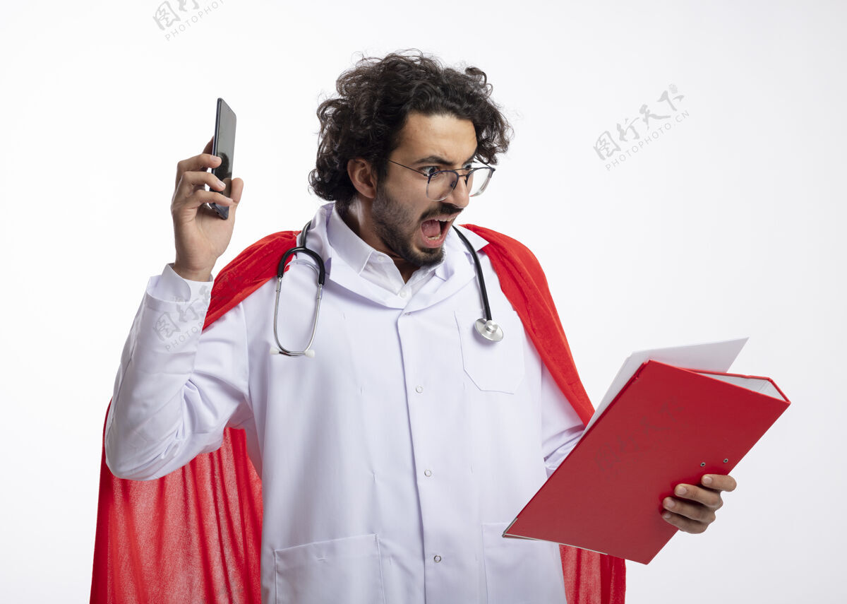 男人一个戴着眼镜 身穿医生制服 披着红色斗篷 脖子上戴着听诊器的年轻白人超级英雄拿着电话 看着白墙上的文件夹制服听诊器斗篷