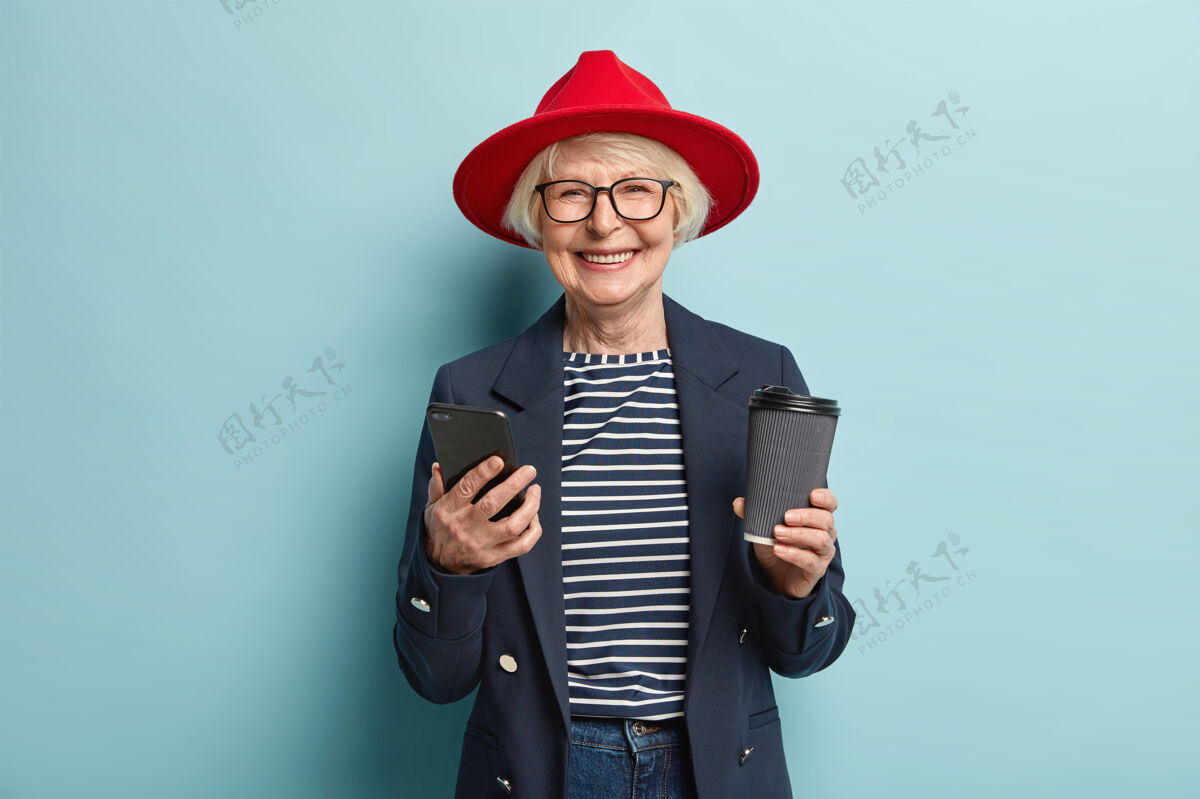 保持戴着眼镜的正面红发美女 使用手机 通过多媒体应用程序发送信息 浏览社交网络 有咖啡休息时间 拿着一次性饮料杯 孤立在蓝色上老人智能手机检查