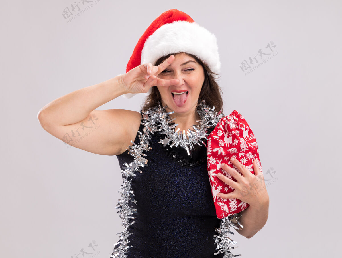 眨眼顽皮的中年妇女戴着圣诞帽 脖子上戴着金属丝花环 手里拿着圣诞礼物袋 看着镜头 舌头和v字符号靠近眼睛 在白色背景上眨眼嬉戏帽子袋子