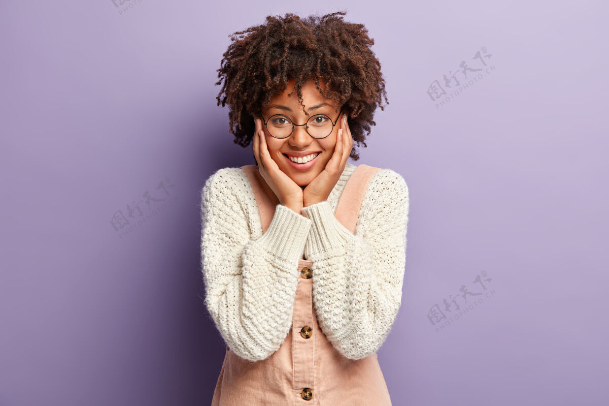 快乐快乐情感概念年轻女性 非洲式发型 双手贴在脸颊上 精神饱满 看到小宝宝很满足 想和孩子一起玩 穿白色针织套头衫和工作服眼镜积极大笑
