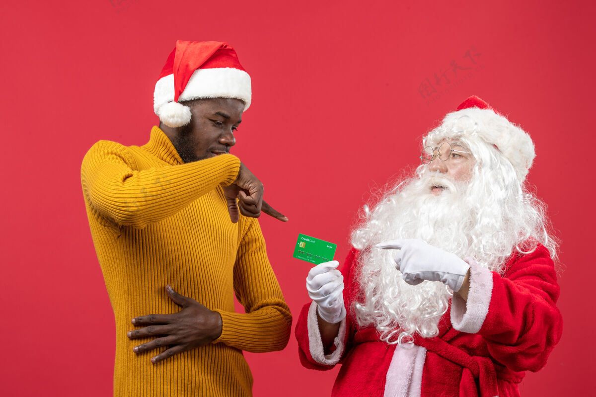 圣诞老人红墙上的圣诞老人和拿着银行卡的年轻人的正视图庆祝圣诞快乐圣诞