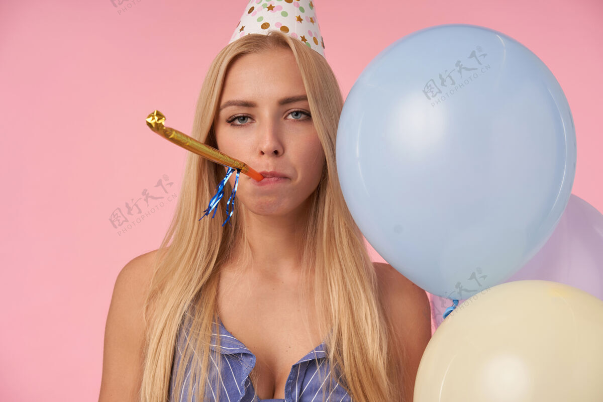 女士毫无灵感的年轻长发金发女性在室内摆好姿势 手里拿着五颜六色的氦气球 吹着派对喇叭 无聊地看着摄像机 站在粉色背景上年轻锥形吹